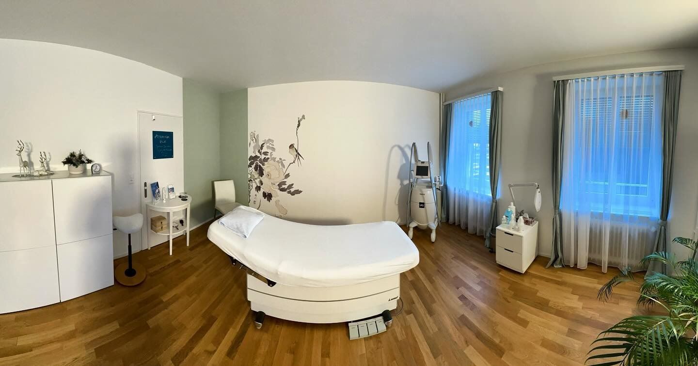 Zimmer mit medizinischer Ausrüstung und einer Behandlungsliege. Weisse und grüne Wände, Holzboden und Fenster.