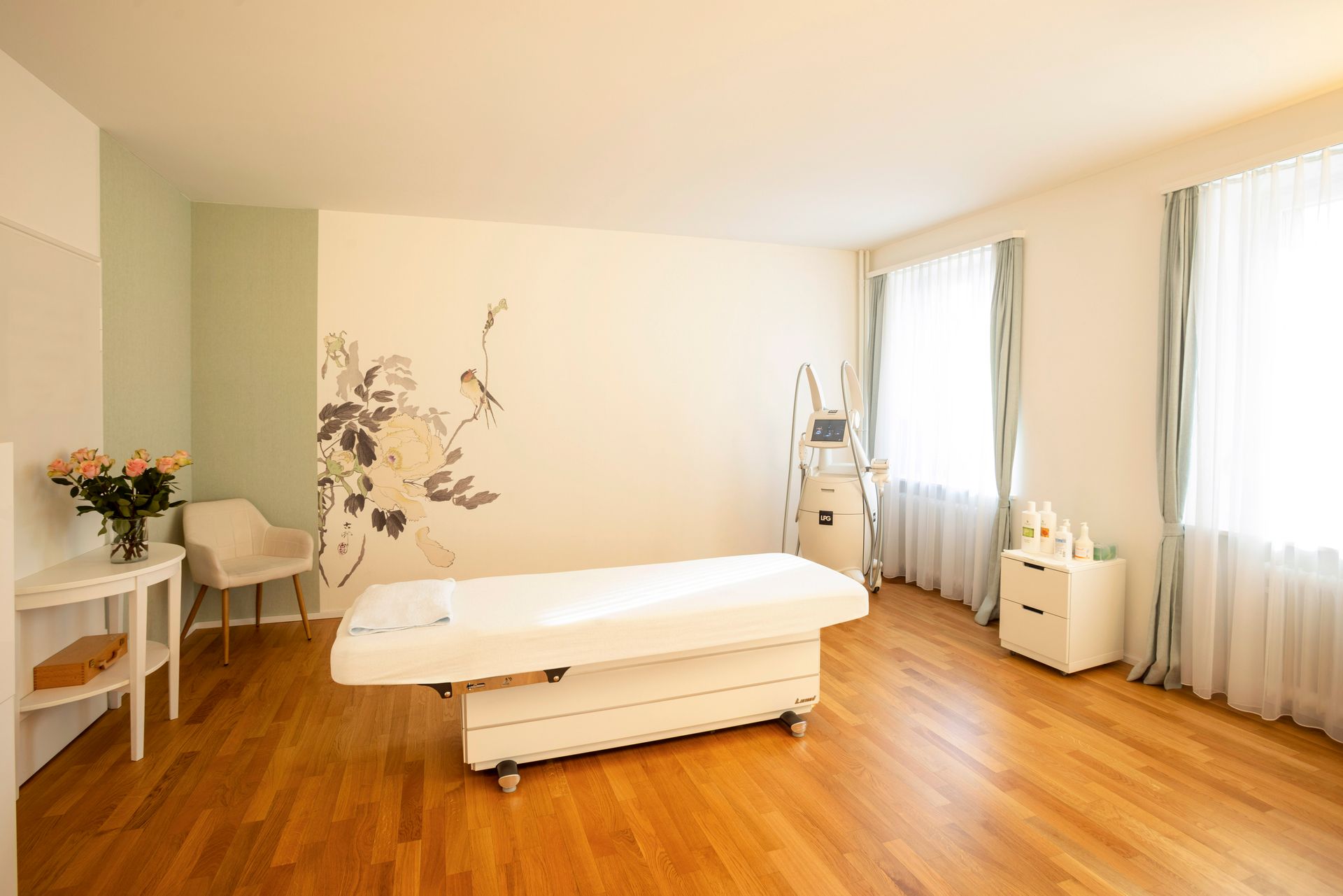 Zimmer mit medizinischer Ausrüstung und einer Behandlungsliege. Weisse und grüne Wände, Holzboden und Fenster.