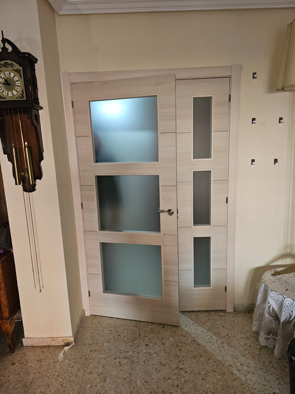 Puerta interior de madera clara con paneles de vidrio esmerilado, junto a un marco a juego. A la izquierda hay un reloj de pie.