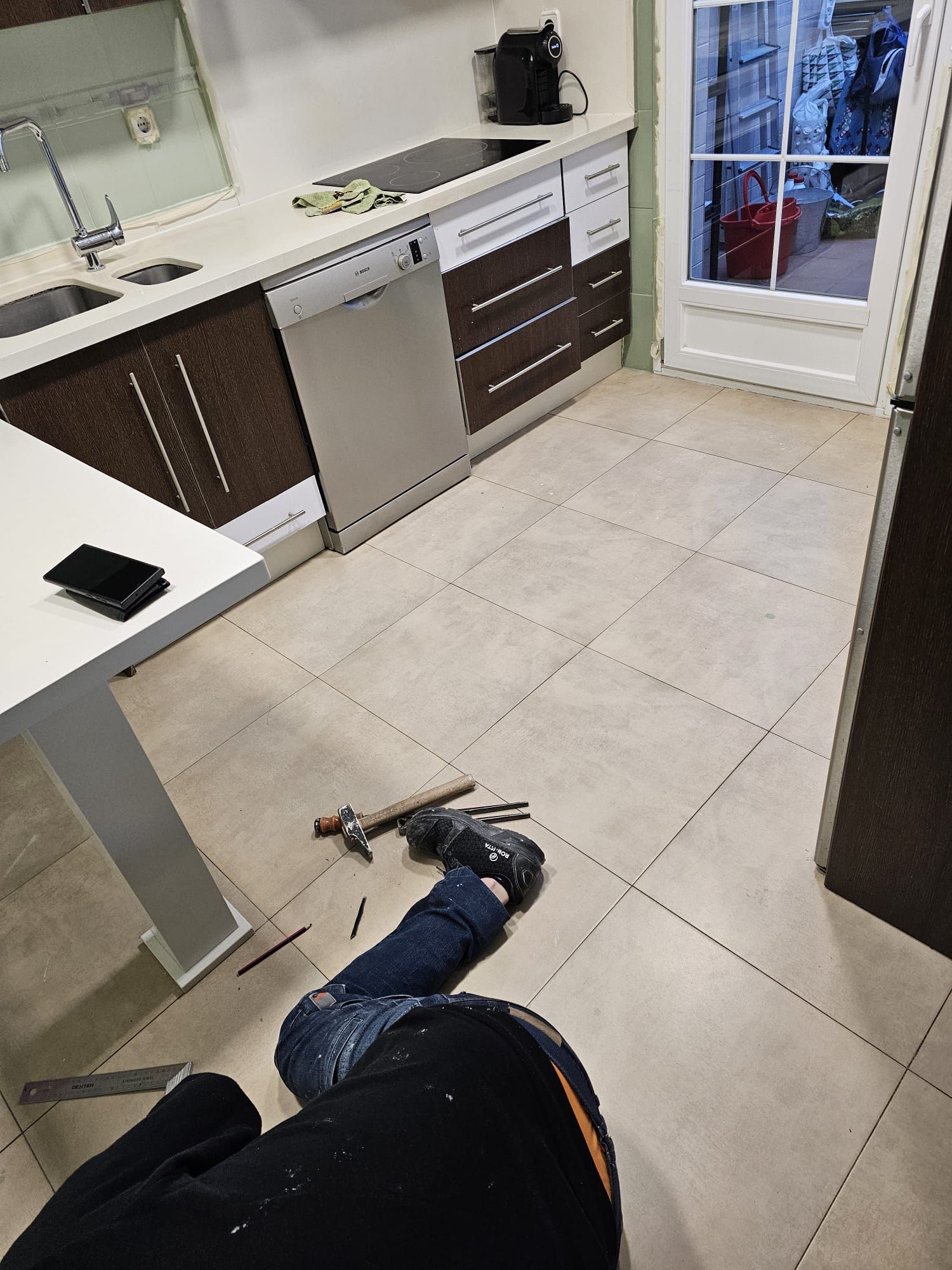 Persona sentada en el suelo de la cocina con herramientas. La cocina tiene encimeras blancas, armarios marrones y lavavajillas de acero inoxidable.