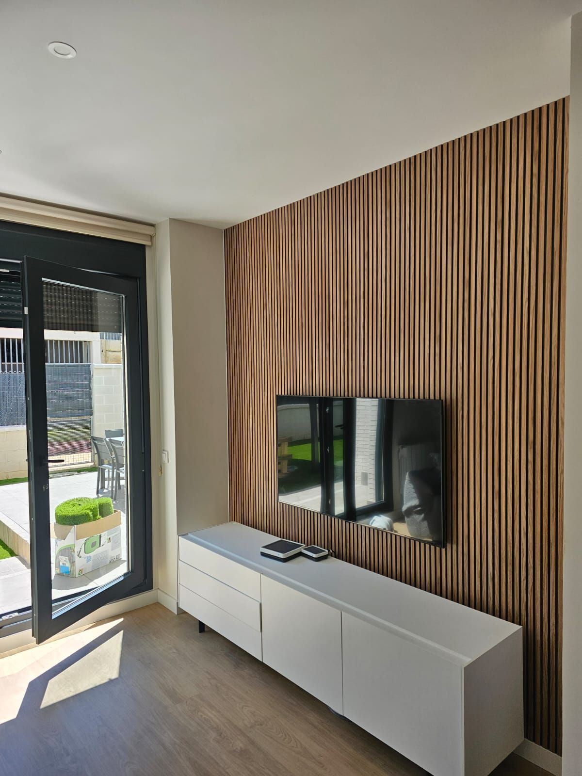 Sala de estar con TV en una pared revestida de madera sobre un mueble blanco; una puerta corrediza de vidrio conduce a un jardín.