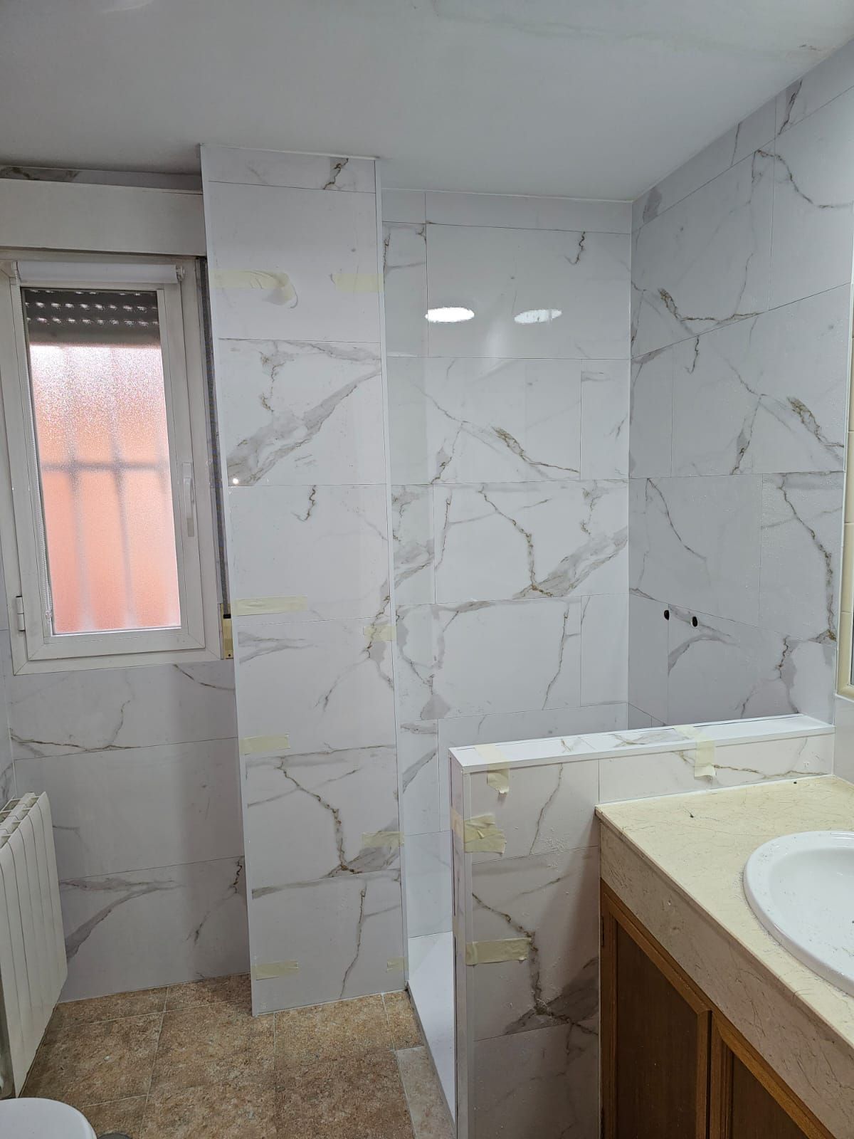 Baño con azulejos de apariencia mármol blanco, una pequeña ventana y un tocador de madera.