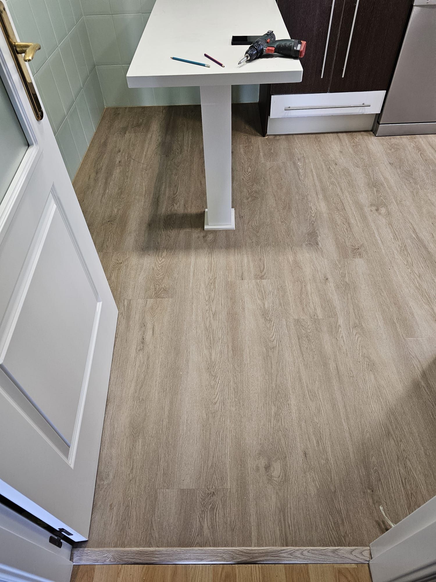 Suelo laminado en una cocina. Mesa blanca con herramientas encima, puerta a la izquierda. Suelo imitación madera clara.