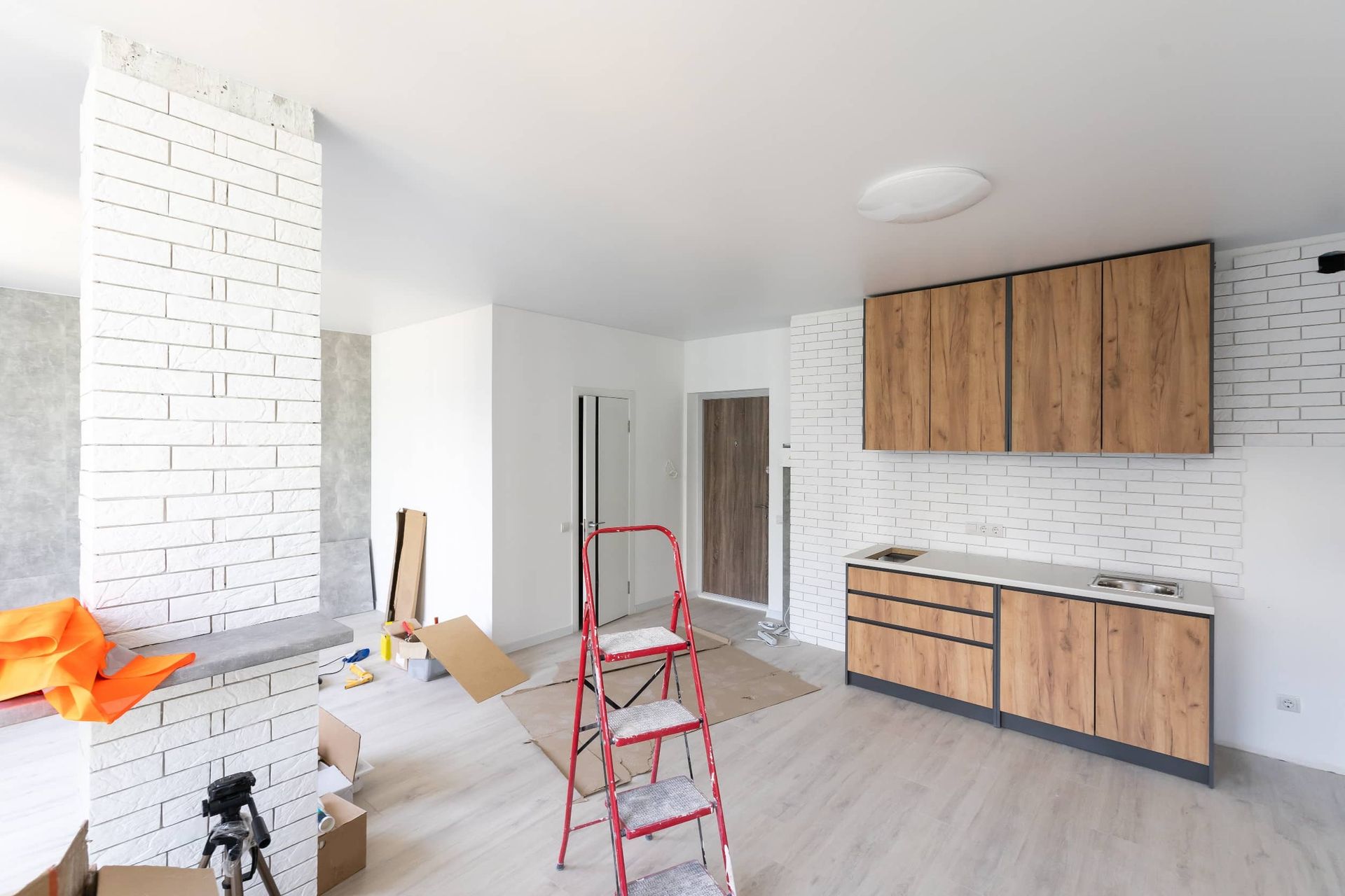 Cocina en remodelación con gabinetes de madera clara, ladrillo blanco, escalera y herramientas.