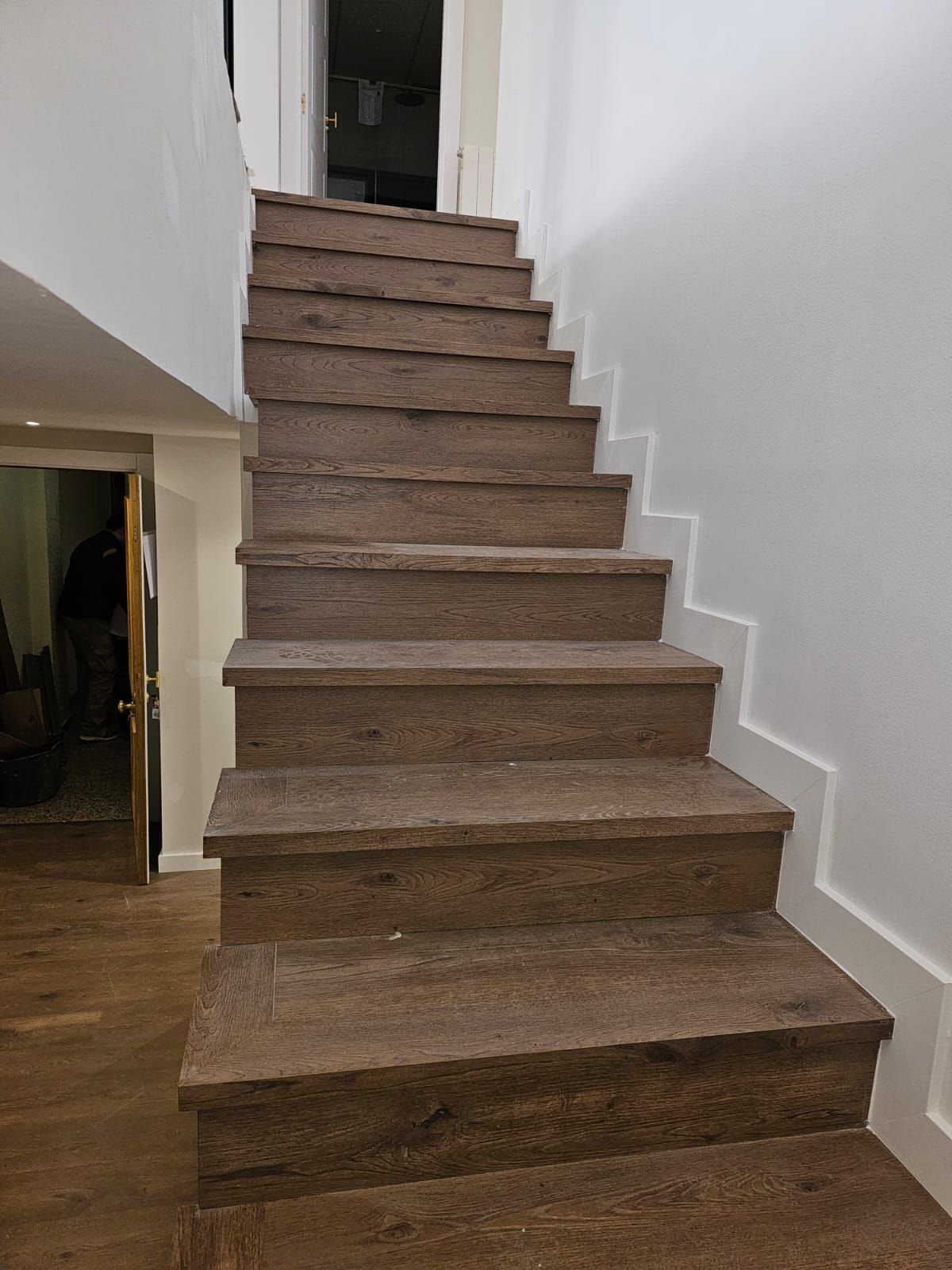 Escalera con peldaños marrones y contrahuellas blancas contra una pared blanca, que conduce hacia arriba, a una puerta.