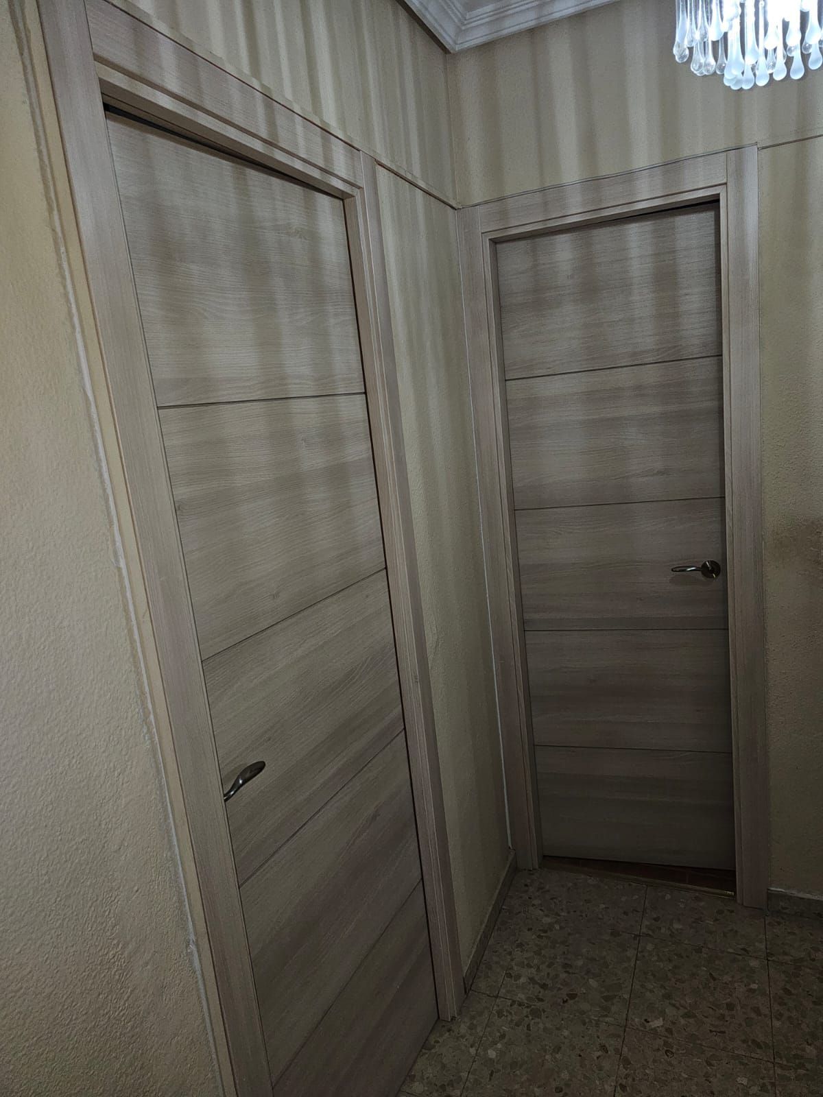 Dos puertas de madera de color claro en un pasillo, una con tirador y otra con pomo; paredes en tonos neutros.