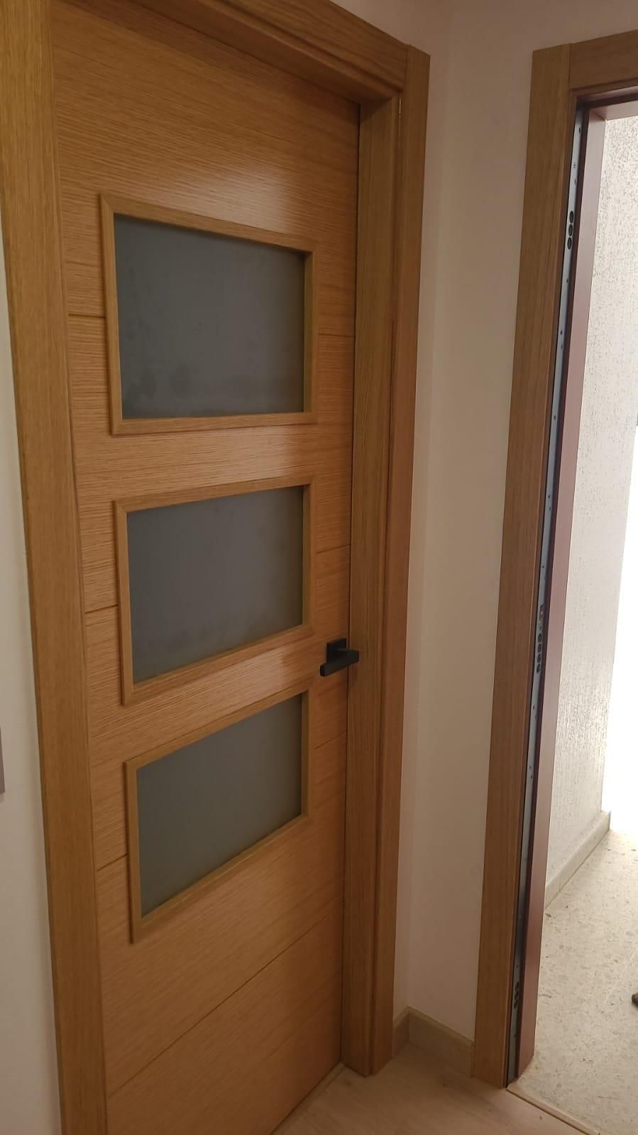 Puerta de madera con tres paneles de vidrio esmerilado, manija negra y marco de madera. Se ve otra puerta.