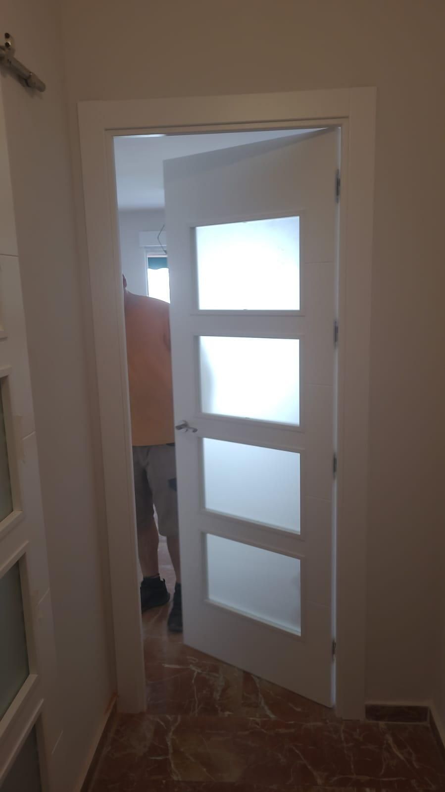 Puerta blanca con paneles de vidrio esmerilado, parcialmente abierta, revelando una persona.