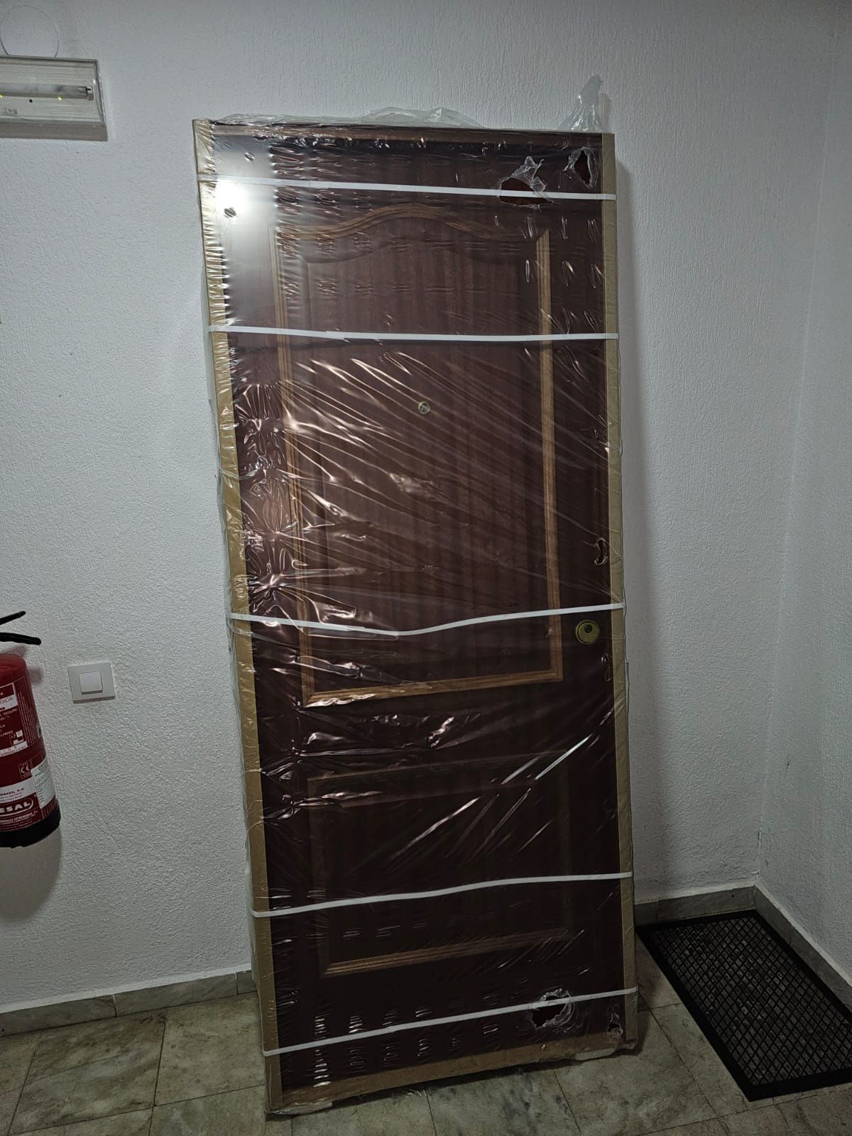 Puerta marrón envuelta apoyada contra una pared blanca; se ve un extintor de incendios.