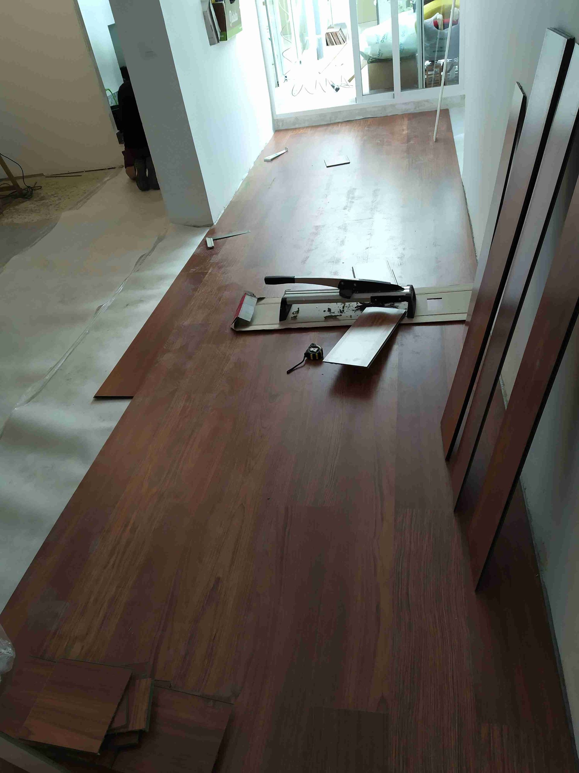 Instalación de pisos de madera en una habitación. Tablas dispuestas, herramientas sobre cartón. Puerta iluminada al fondo.