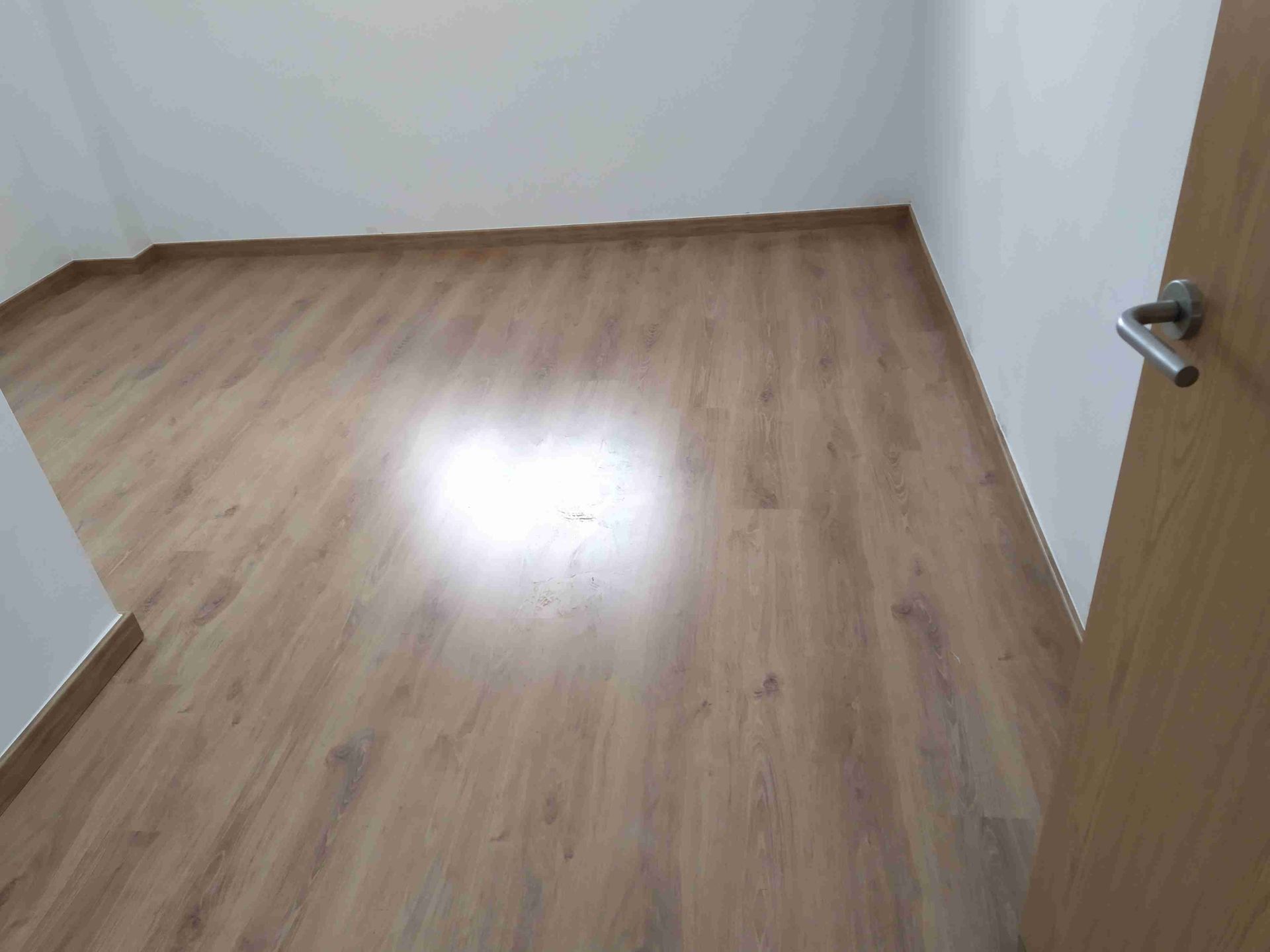 Habitación vacía con suelo imitación madera y paredes blancas. La luz se refleja en el suelo. Puerta a la derecha.