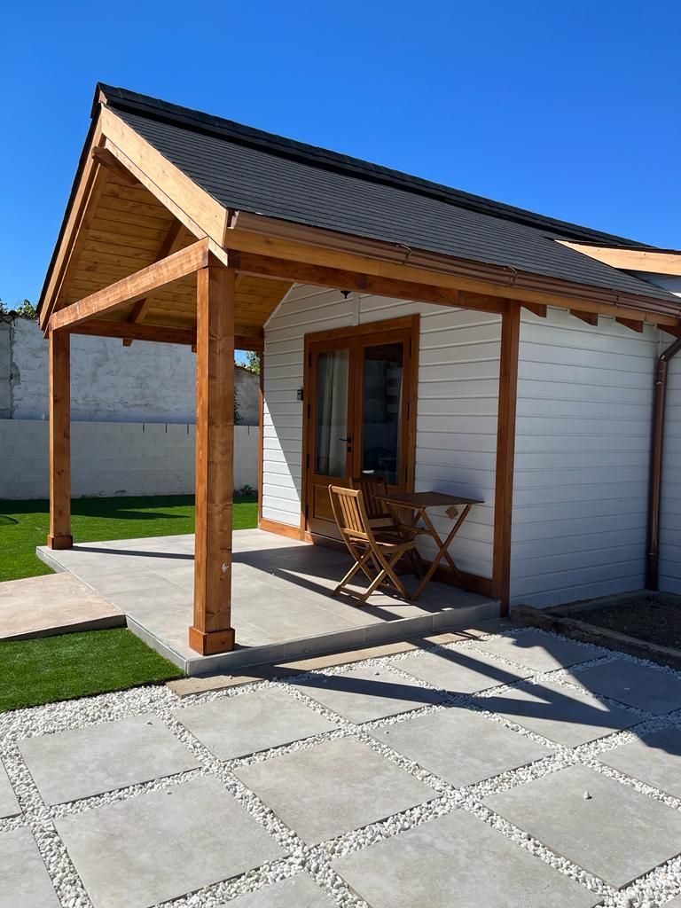 Una pequeña casa blanca con un porche de madera y una mesa y sillas delante.