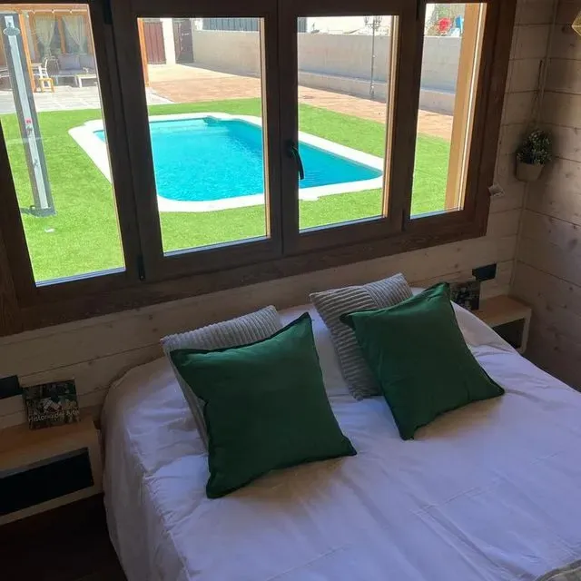 Una cama con almohadas verdes frente a una ventana con una piscina al fondo.