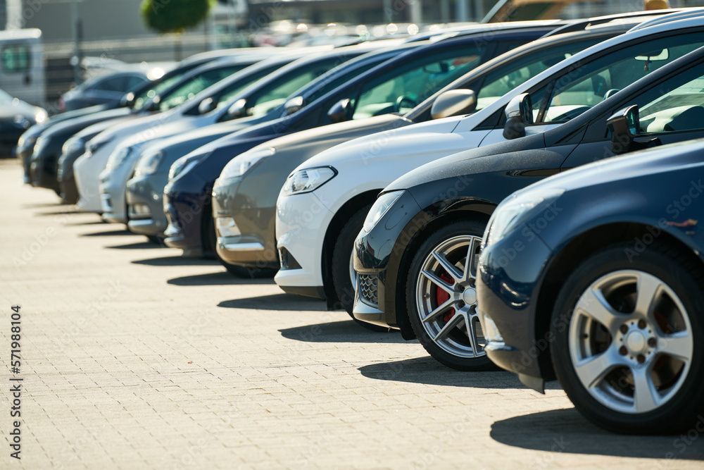 Autos stehen in einer Reihe auf einem Parkplatz im Freien. Verschiedene Farben, darunter Weiss, Blau und Schwarz. Sonniger Tag.