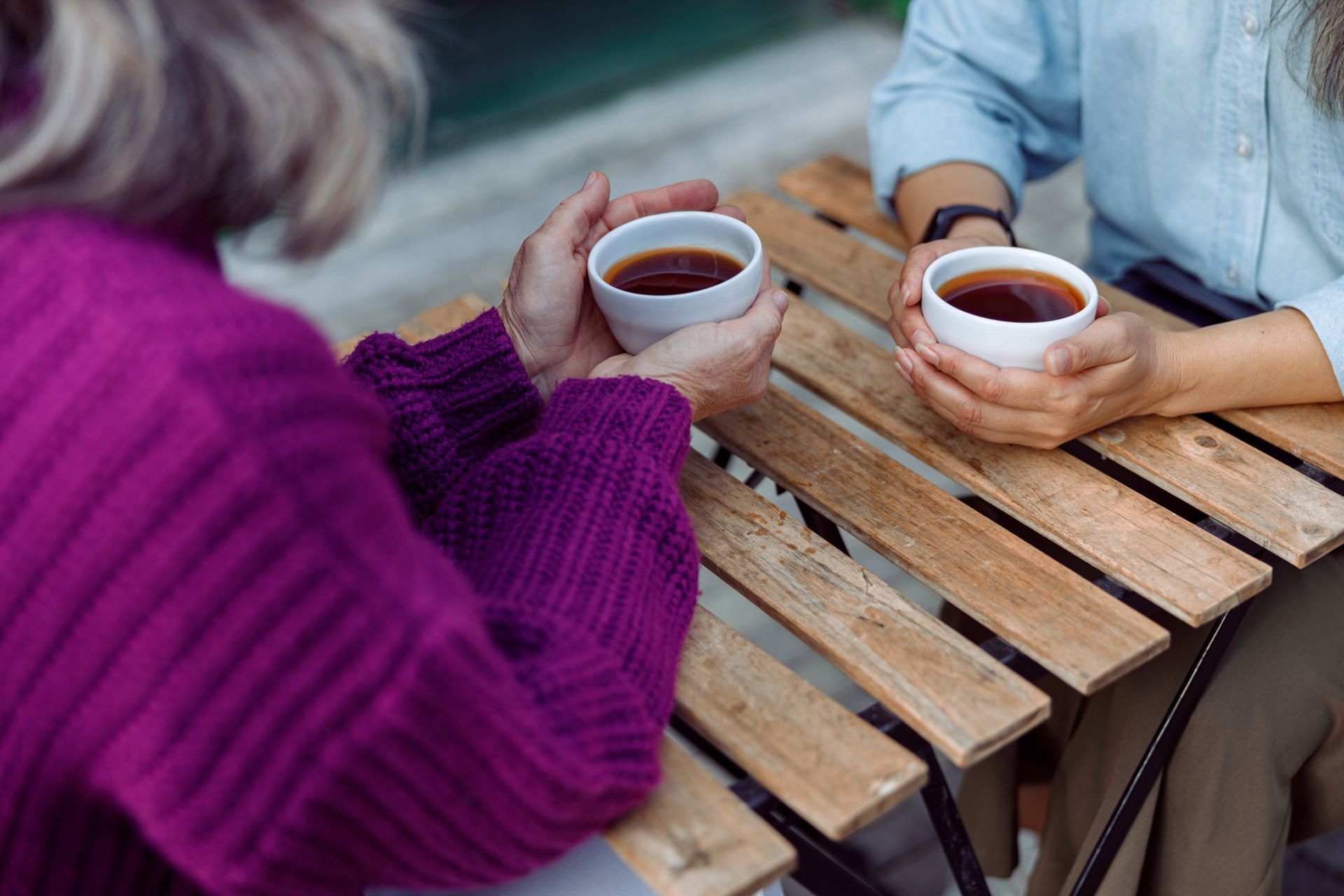 Deux personnes, tasses de thé blanches à la main, sont assises à une table en bois à l'extérieur.