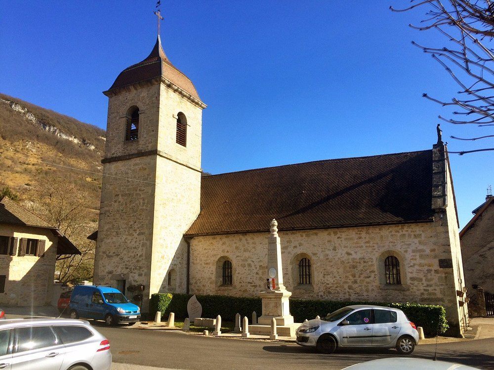 Église en pierre avec clocher et des voitures garées dans la rue, montagnes en arrière-plan.