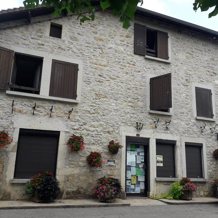Bâtiment à Montagnieu en pierre aux volets et portes marron, orné de paniers de fleurs.