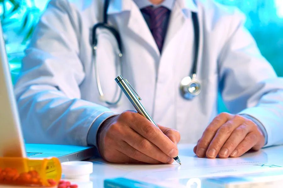 Un médico está sentado en un escritorio escribiendo en un trozo de papel.