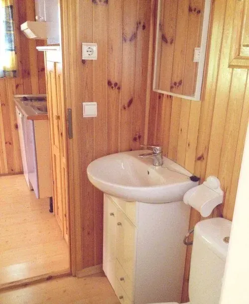 Baño pequeño con paneles de madera, lavabo blanco y espejo junto al inodoro.