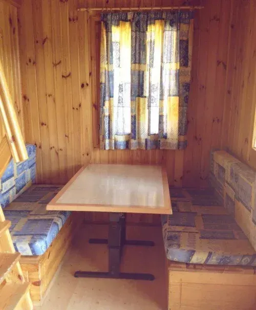 Rincón de cabaña con paneles de madera, bancos acolchados, una mesita y una ventana con cortinas.