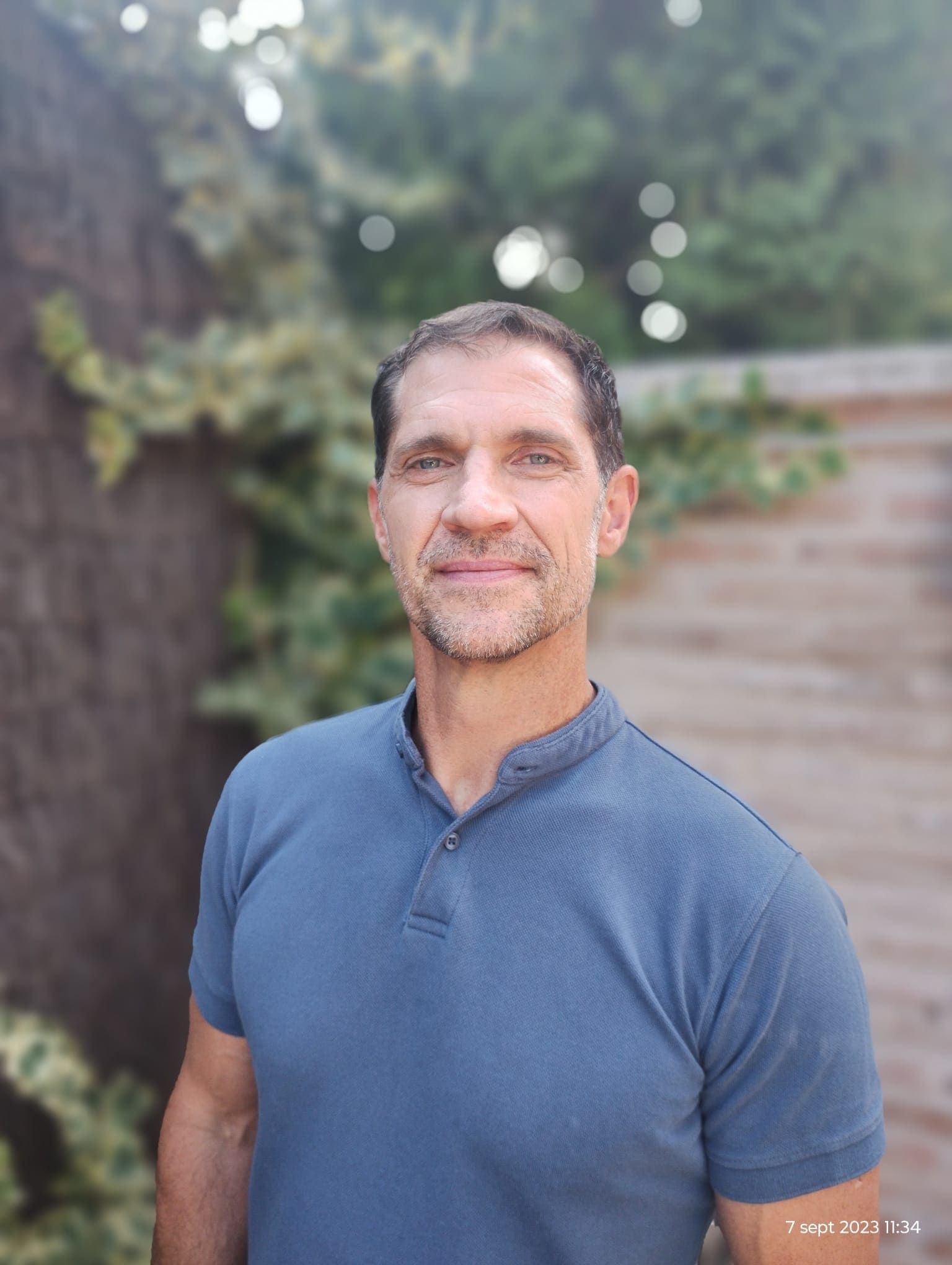 Un hombre con polo azul sonríe en el exterior, con vegetación al fondo.