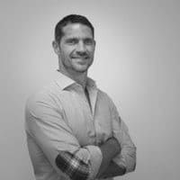 Hombre con camisa abotonada y coderas de cuadros, brazos cruzados, sonriendo.