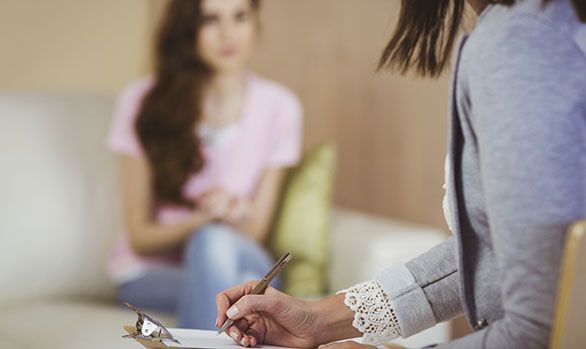 Una mujer está sentada en un sofá hablando con una psicóloga que está escribiendo en un portapapeles.