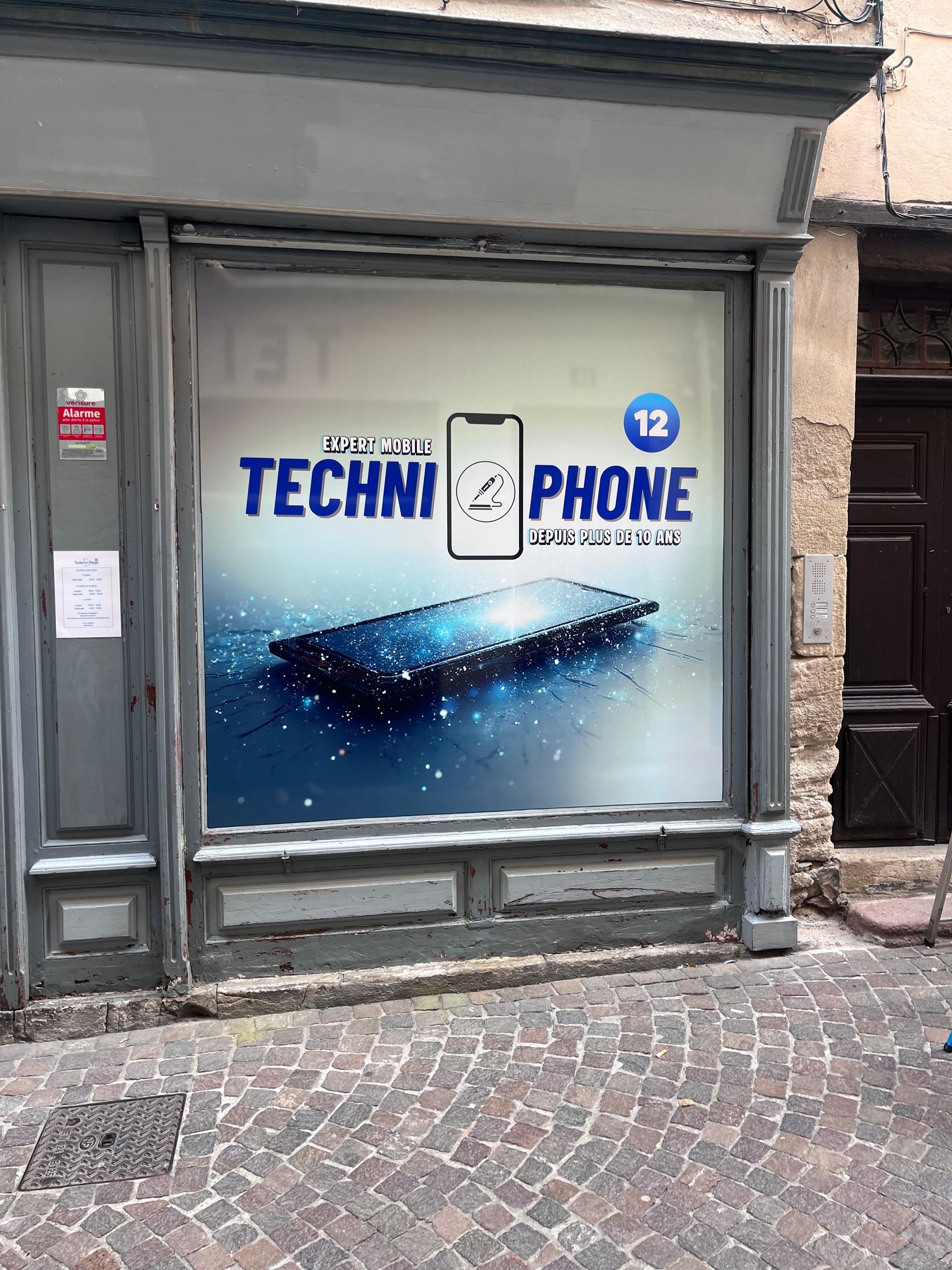 Une devanture de magasin avec une grande publicité en vitrine pour  TECHNIPHONE  présentant un dessin de smartphone bleu et un emblème 12 .