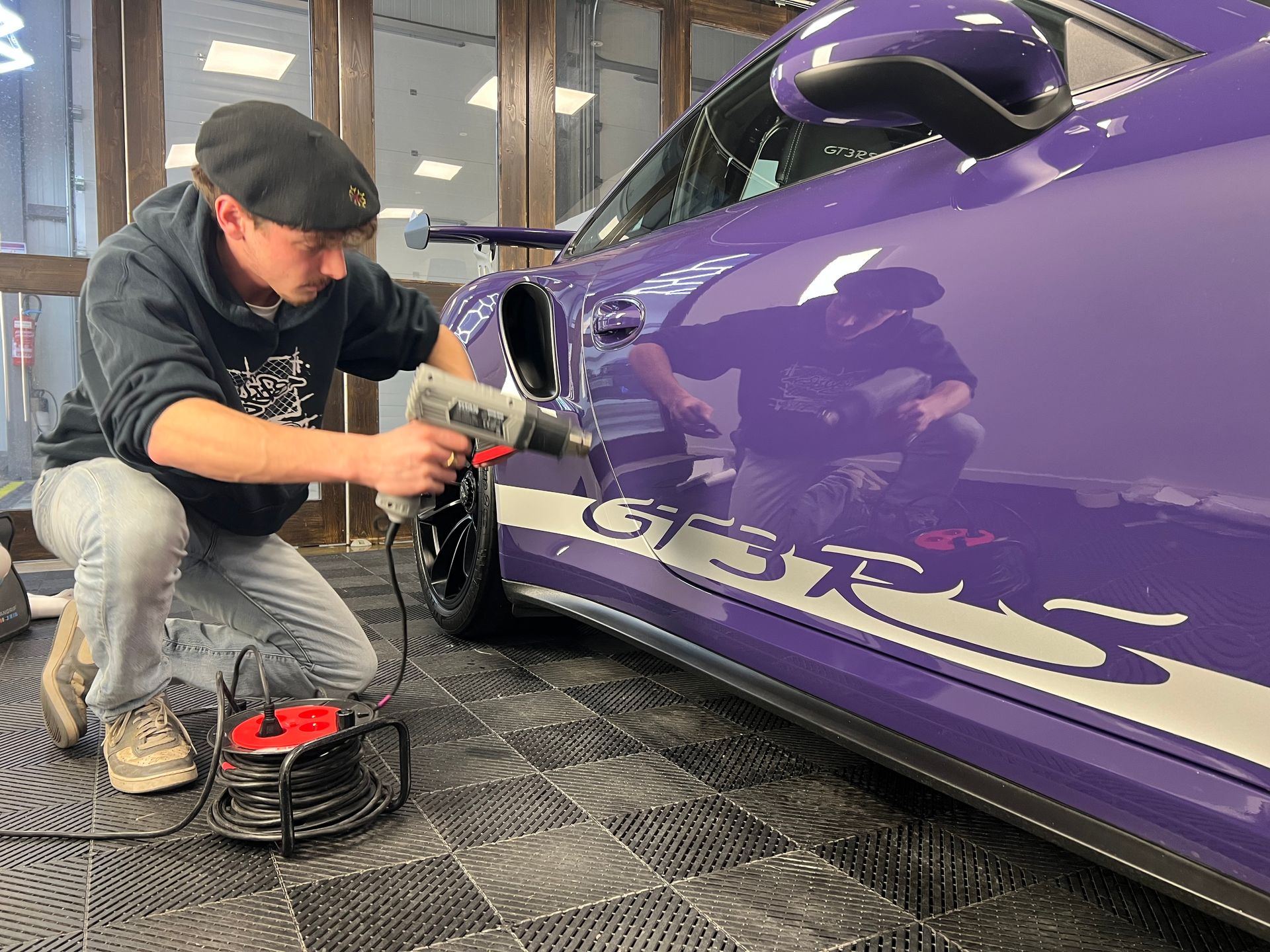 Dans un atelier, une personne utilise un pistolet thermique pour appliquer un autocollant blanc GT3 RS sur le côté d'une voiture de sport violette.