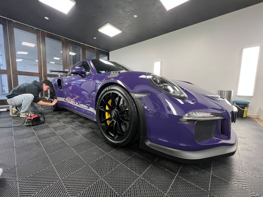 Une personne installe un kit adhésif sur une Porsche violette dans un studio de detailing automobile professionnel et lumineux.