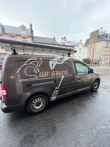 Une camionnette de service marron garée dans une rue mouillée, avec une échelle de toit et des graphismes blancs représentant une clé et un marteau.