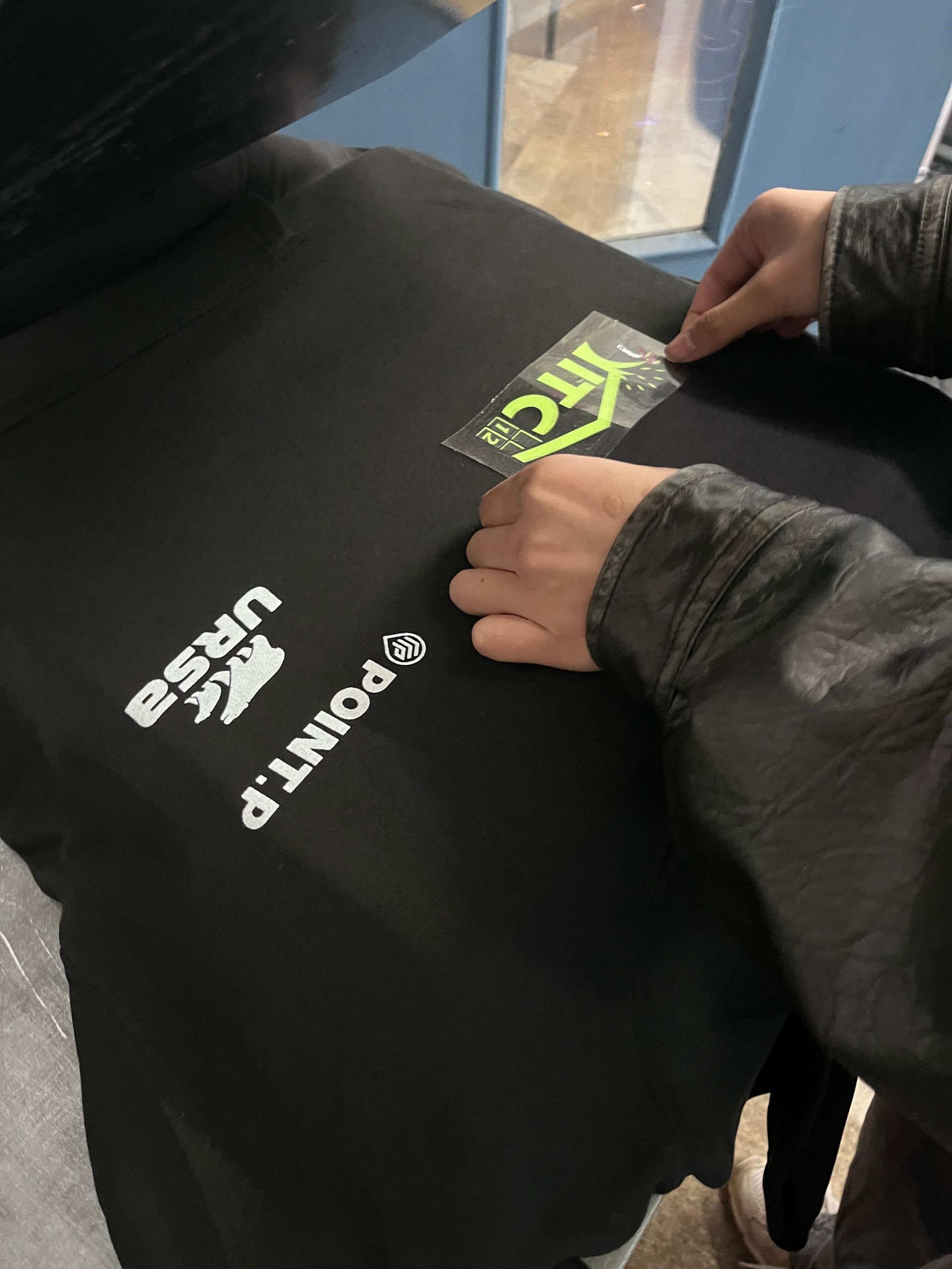Une personne aligne un autocollant thermocollant jaune fluo  ITC sur un t-shirt noir comportant déjà des logos d'entreprise blancs.