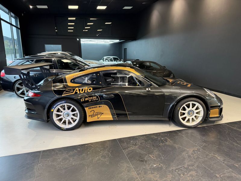 Une Porsche GT3 de course noire et or est garée dans un showroom moderne et bien éclairé, avec d'autres véhicules en arrière-plan.