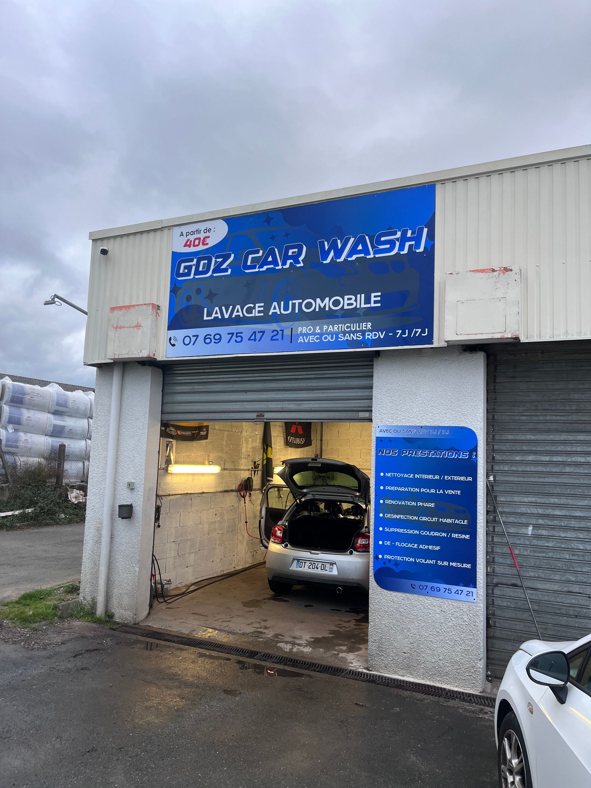 Extérieur du lave-auto Goz avec une enseigne bleue montrant une voiture argentée dans l'aire de service et une liste de prix affichée au mur.