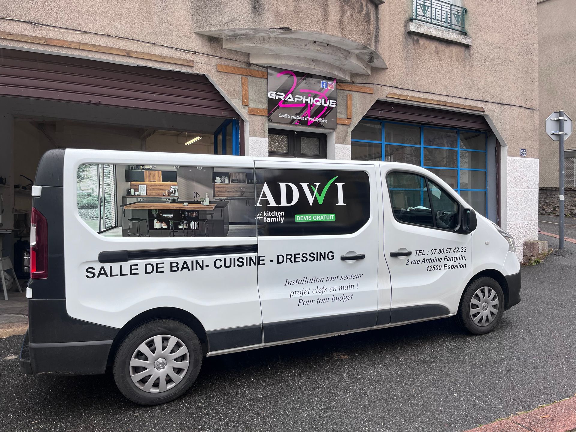 Une camionnette blanche de la société ADWI avec l'inscription SALLE DE BAIN - CUISINE - DRESSING sur le côté, garée devant un magasin.
