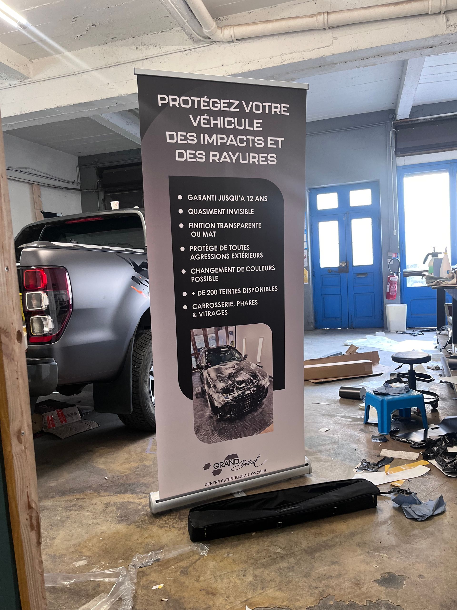 Une banderole publicitaire verticale, installée dans un garage à côté d'un camion gris mat, fait la publicité de services de protection de véhicules.