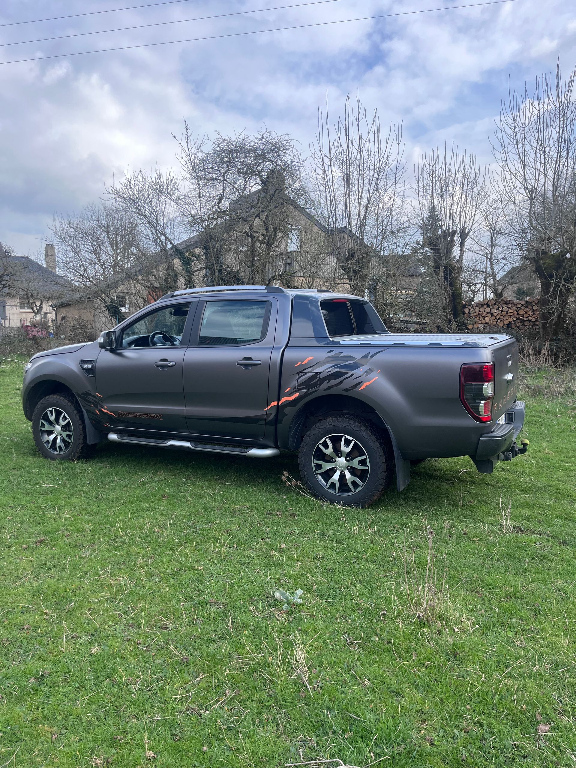 Un pick-up gris foncé est garé sur un terrain herbeux, devant des arbres dénudés et un bâtiment.