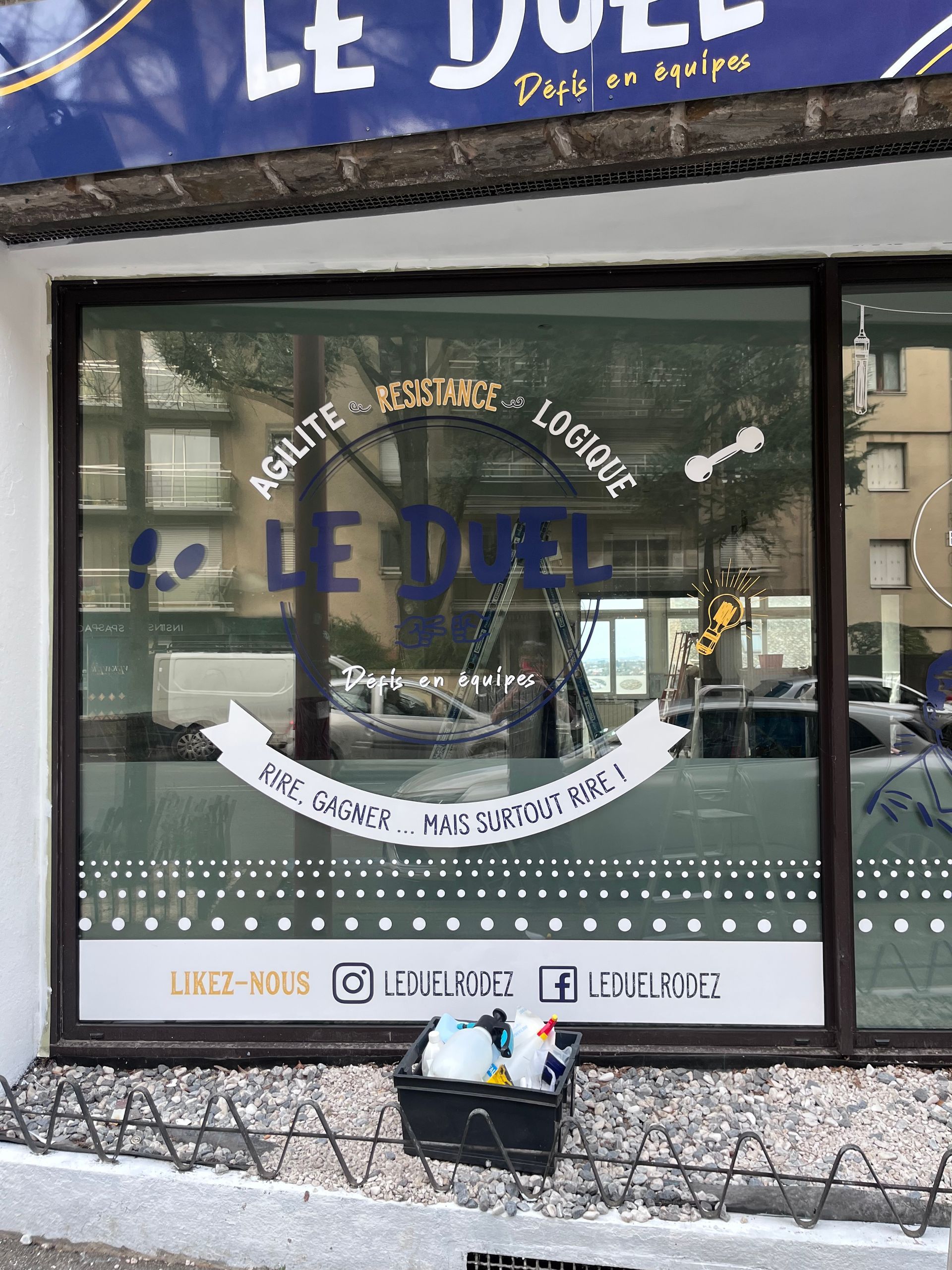 La façade d'un commerce appelé LE DUEL arbore des icônes de réseaux sociaux et du texte en français sur la vitrine.