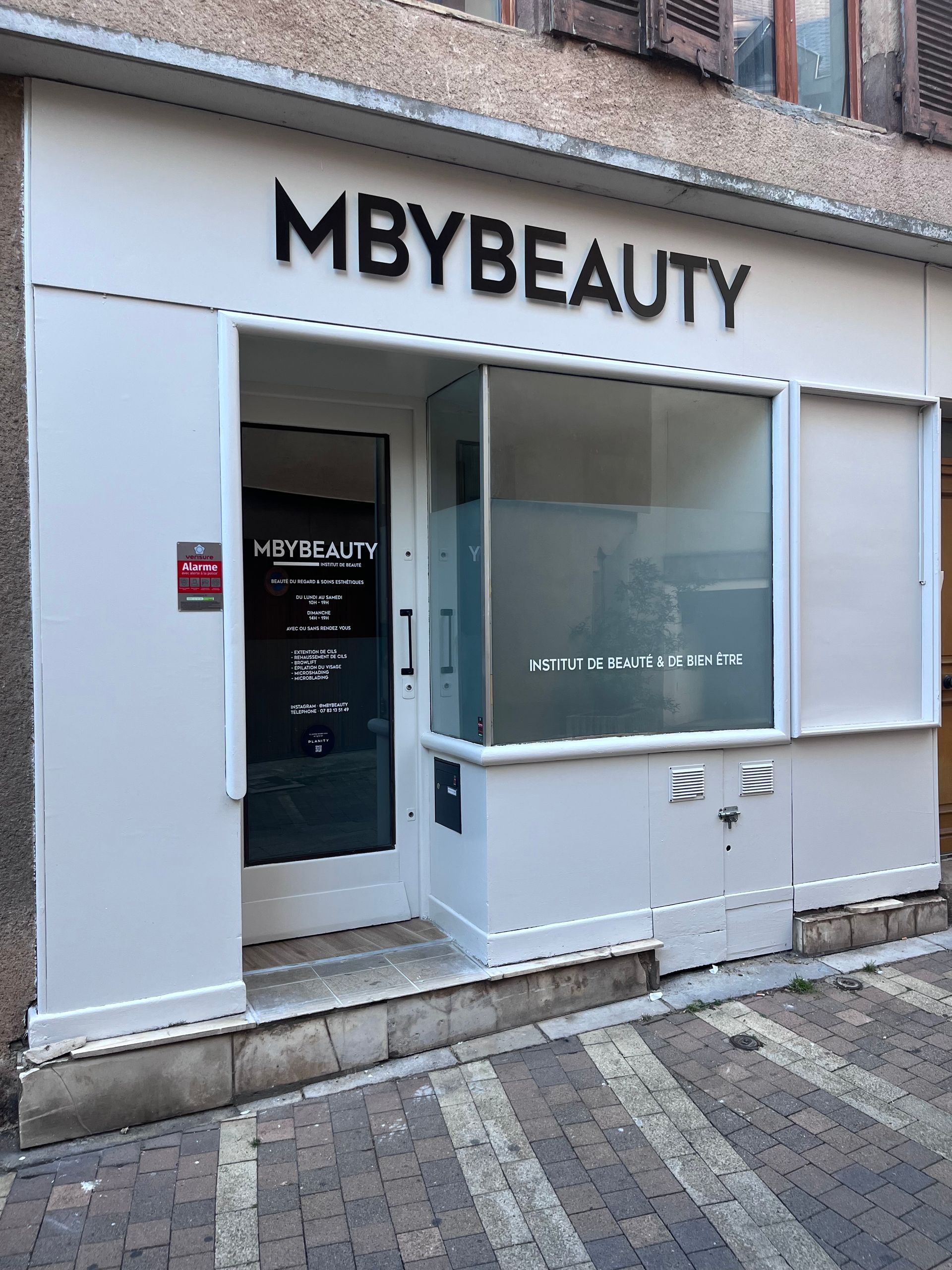 Une façade pour l'enseigne MBYBEAUTY, avec des murs blancs, une grande vitrine et une porte vitrée, située dans une rue pavée.