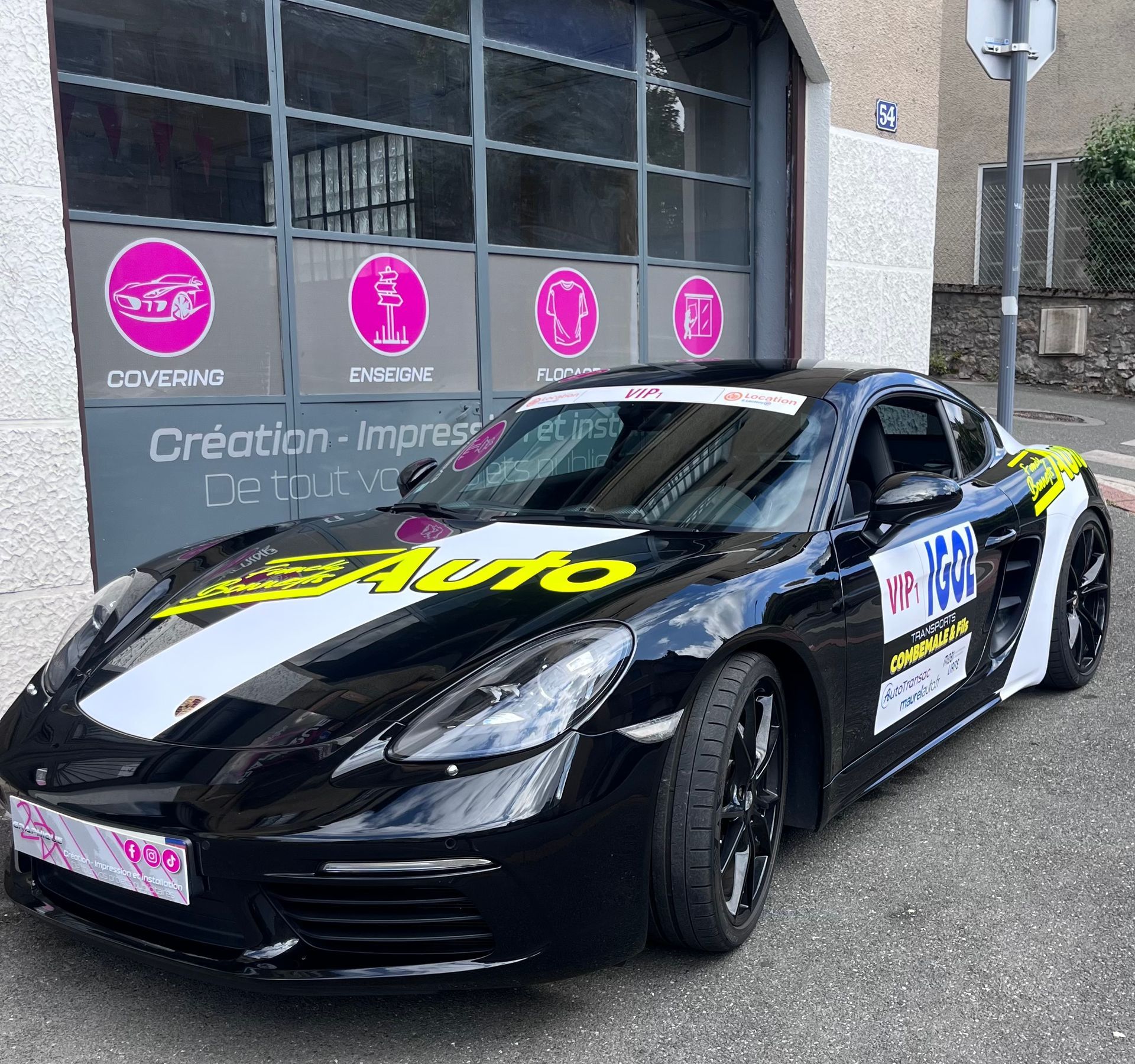 Une Porsche noire, voiture de sport arborant un logo publicitaire, est garée devant un magasin dont la vitrine présente des icônes de service roses.