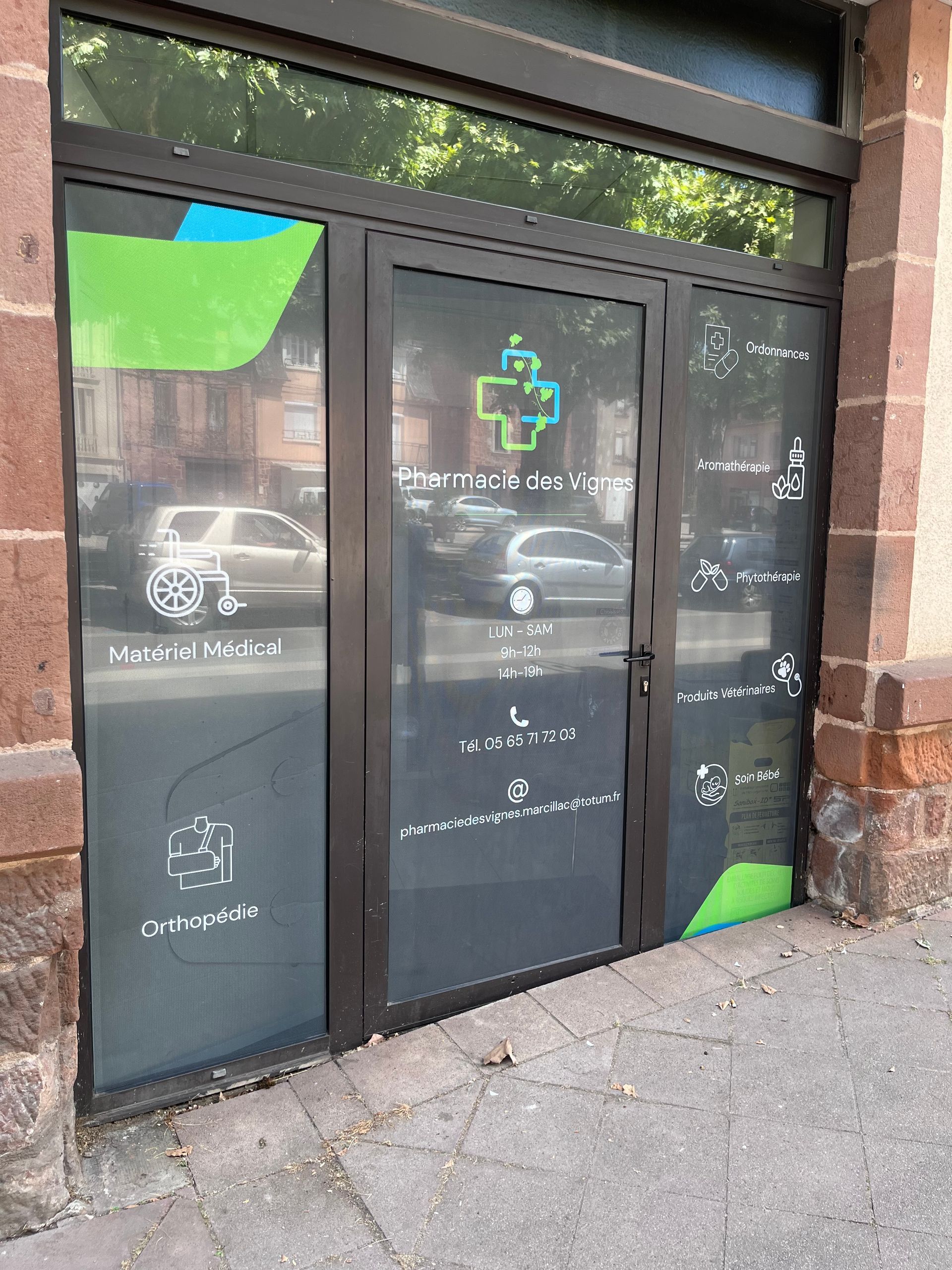 L'entrée d'une pharmacie avec des portes vitrées, ornée d'un logo en forme de croix verte et bleue et d'icônes blanches sur un fond sombre et semi-opaque.