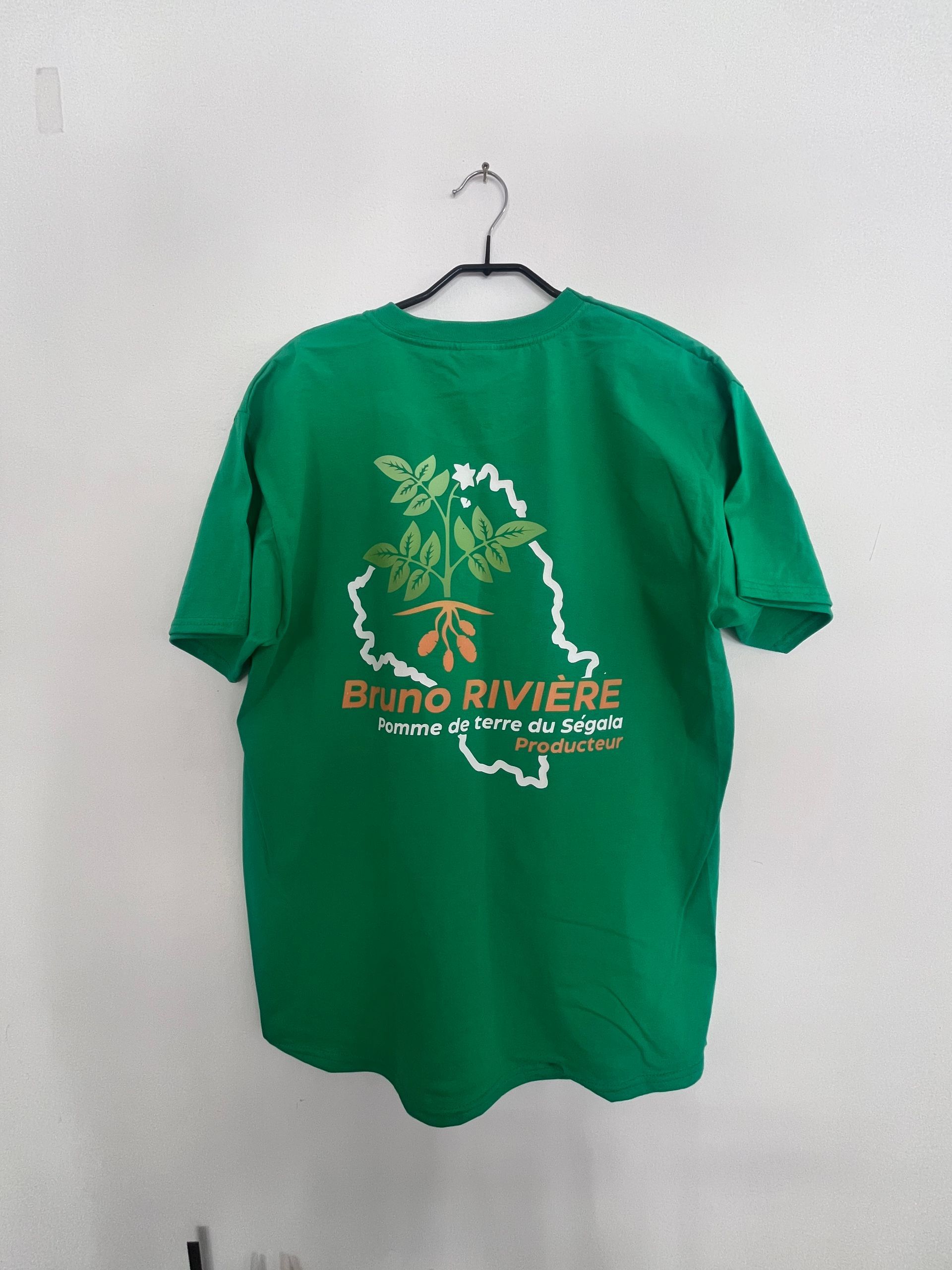 Un t-shirt vert accroché à un cintre, orné d'un contour blanc d'une région, d'un dessin de plante et du texte Bruno RIVIÈRE.