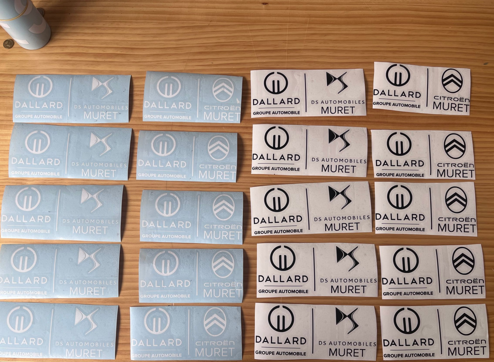 Des rangées d'autocollants en vinyle blanc arborant les logos Dallard, DS et Citroën Muret sont disposées sur une surface en bois.