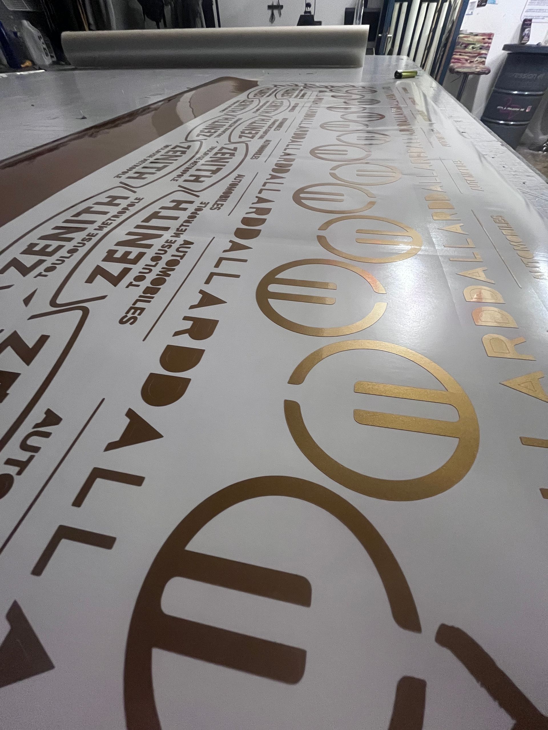 Vue grand angle d'une grande feuille d'autocollants en vinyle comportant des textes et des logos dorés, disposée sur une table d'atelier.
