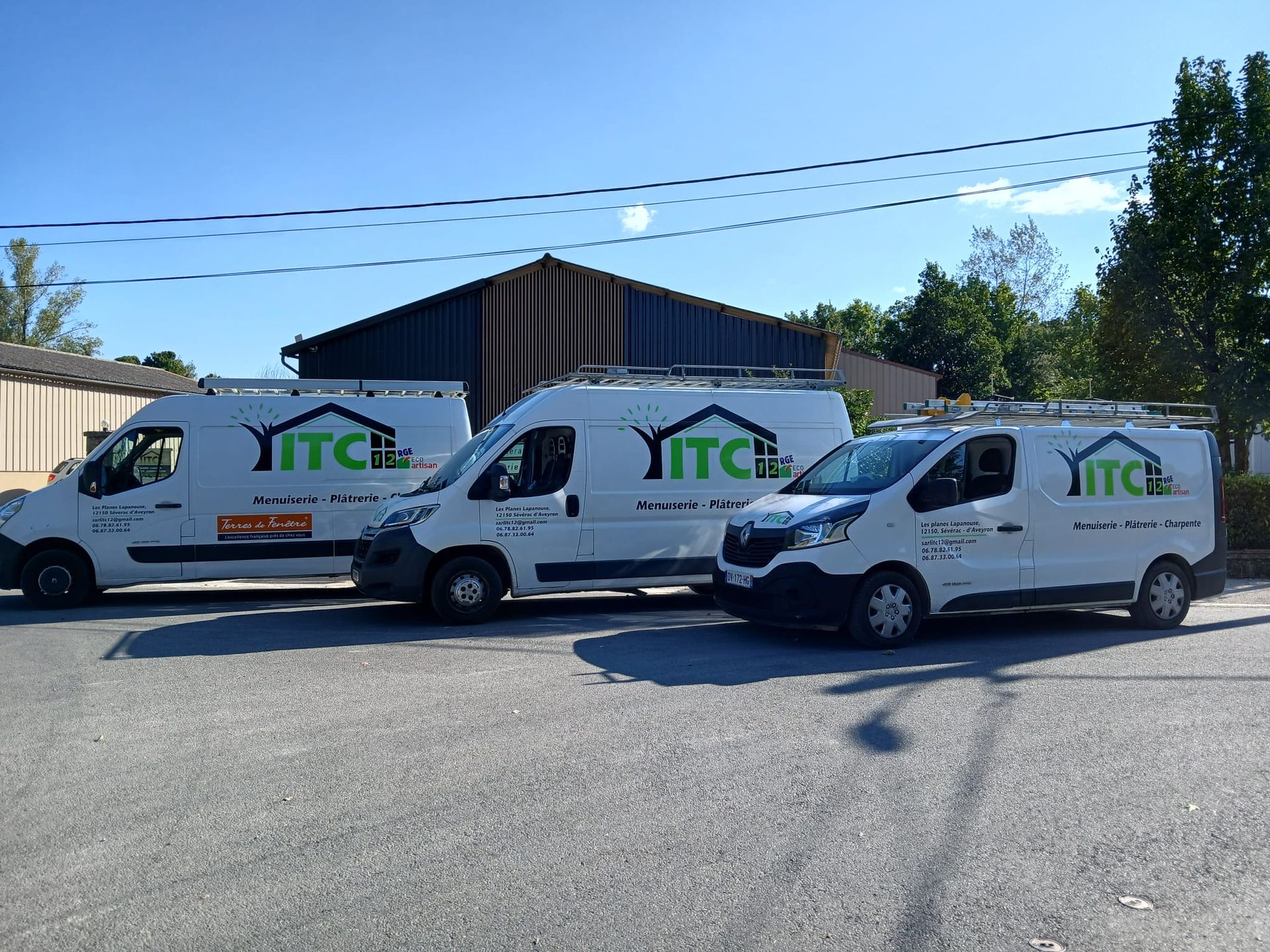 Trois fourgonnettes blanches de chantier portant le logo ITC étaient garées en rang sur un terrain gravillonné sous un ciel bleu azur.
