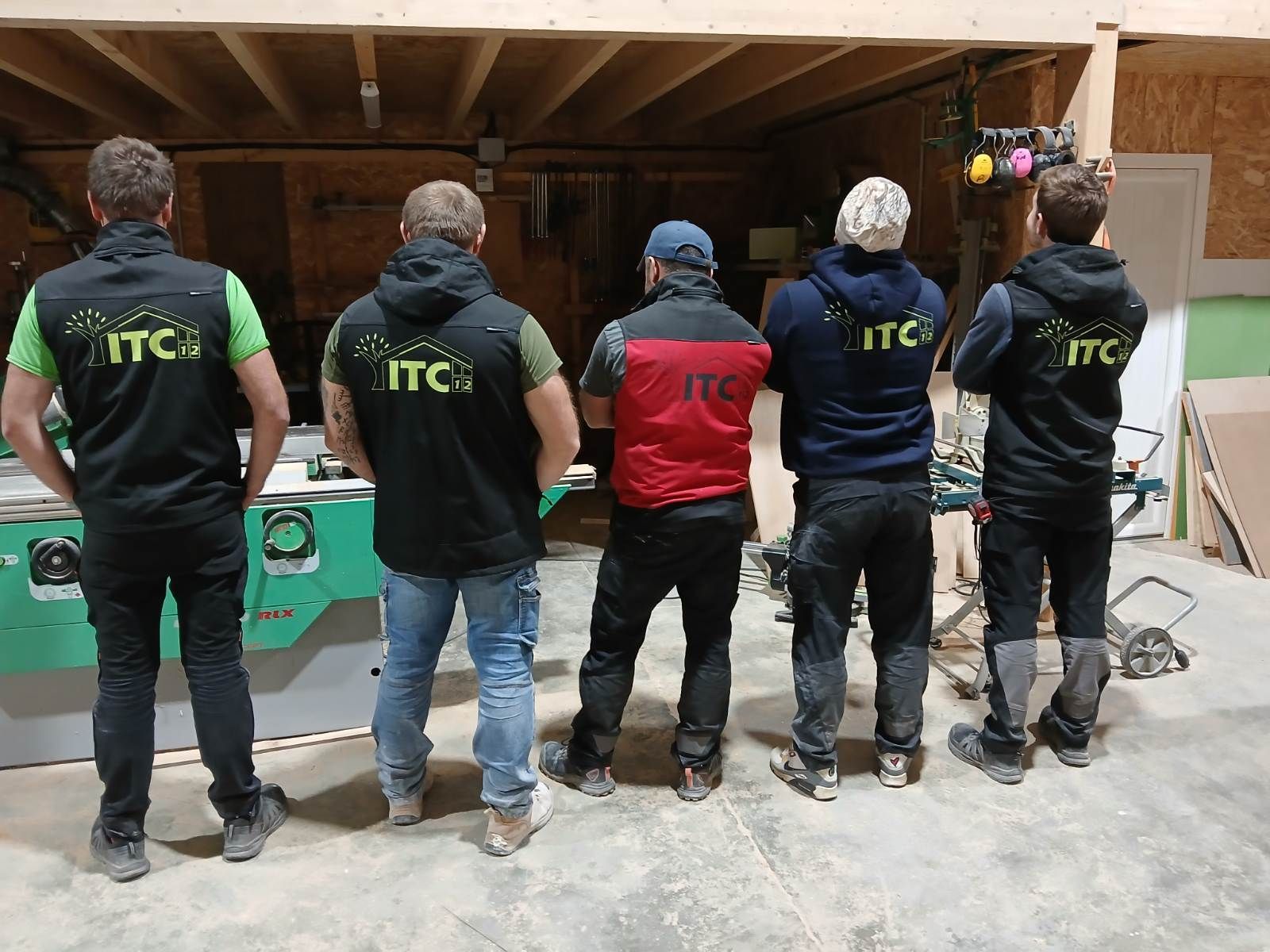Cinq personnes debout en rang, dos à dos, vêtues de vêtements sombres avec des logos ITC, dans un atelier.