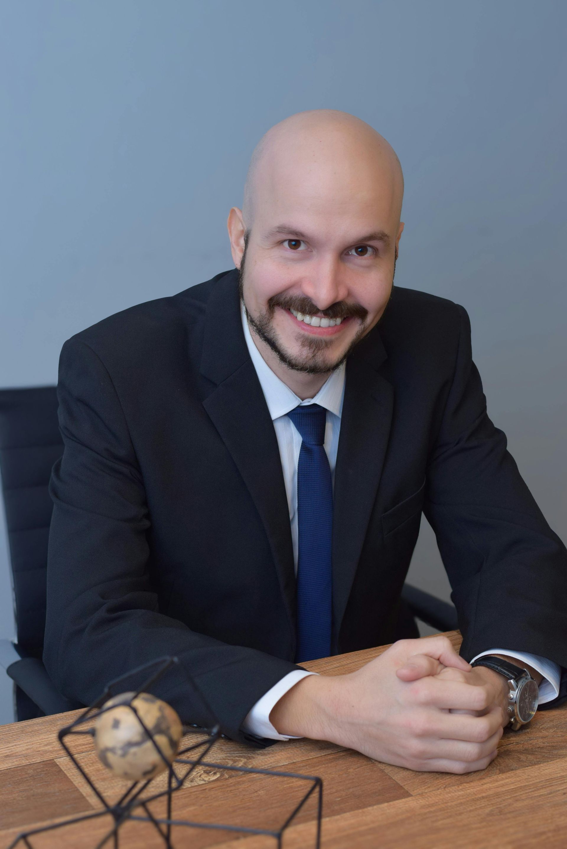 Glatzköpfiger Mann im Anzug lächelt in die Kamera; sitzt am Schreibtisch.