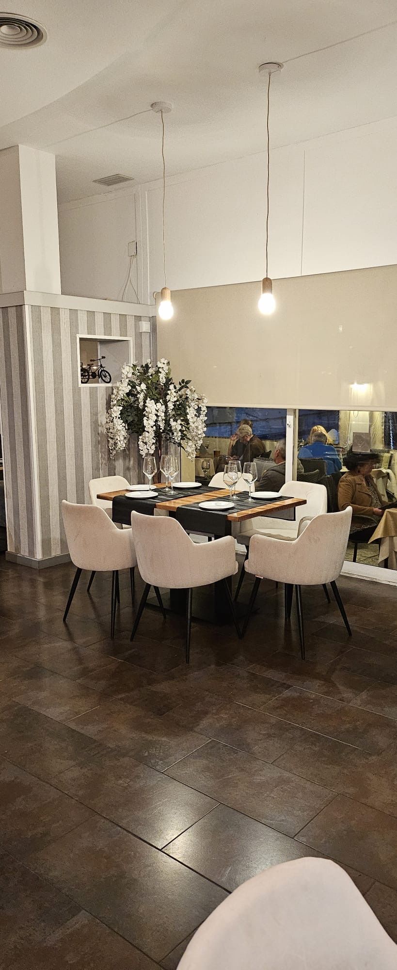 Comedor moderno con sillas blancas, una mesa redonda, lámparas colgantes y suelo de baldosas oscuras.