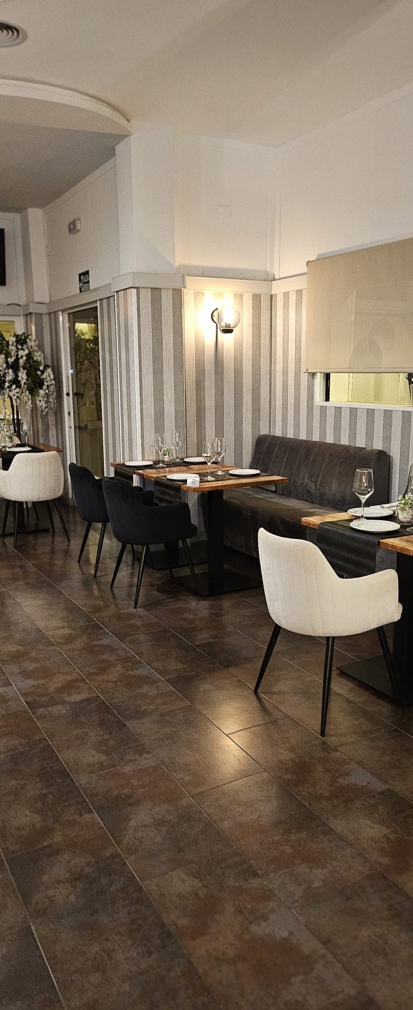 Interior de restaurante moderno con mesas largas, sillas blancas, iluminación cálida y suelo de baldosas oscuras.