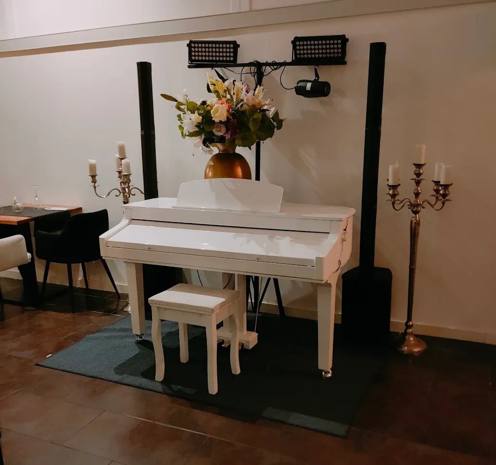 Piano vertical blanco con arreglo floral y un pequeño banco sobre una alfombra en una habitación con iluminación cálida.